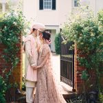 South Asian wedding couple embracing in traditional ceremony attire framed by lush garden gateway and historic home