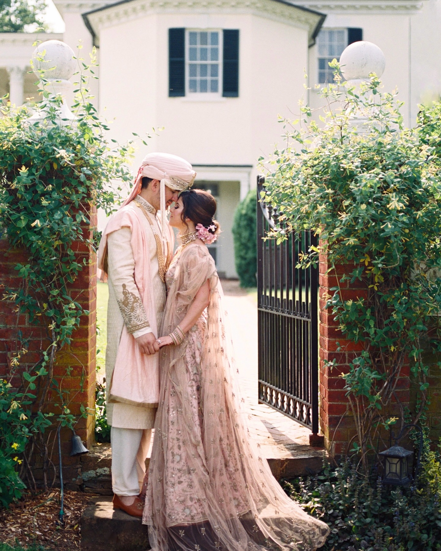 South Asian wedding couple embracing in traditional ceremony attire framed by lush garden gateway and historic home