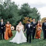 Bride and groom pose with wedding party in orange and burgundy attire on grassy lawn under cloudy sky