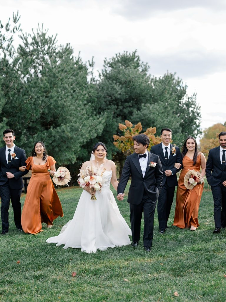 Bride and groom pose with wedding party in orange and burgundy attire on grassy lawn under cloudy sky