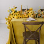 Sweetheart table with mustard yellow linen, rustic wooden chairs, and cascading yellow dahlia centerpiece with gold taper candle