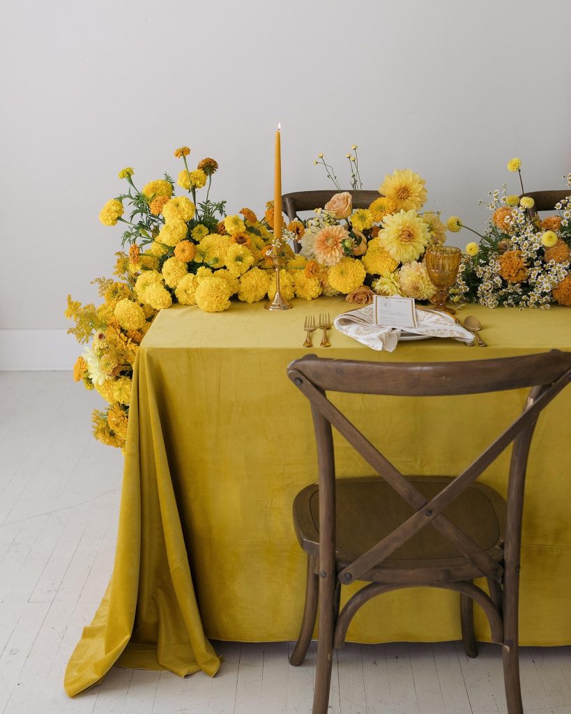 Sweetheart table with mustard yellow linen, rustic wooden chairs, and cascading yellow dahlia centerpiece with gold taper candle