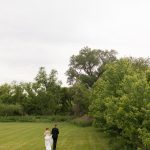 Bride and groom walk away across manicured lawn bordered by lush green trees and natural landscape