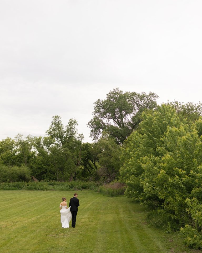 Bride and groom walk away across manicured lawn bordered by lush green trees and natural landscape