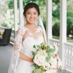 Bride in lace wedding dress with illusion neckline and veil, wearing updo hairstyle, holding white and greenery bouquet