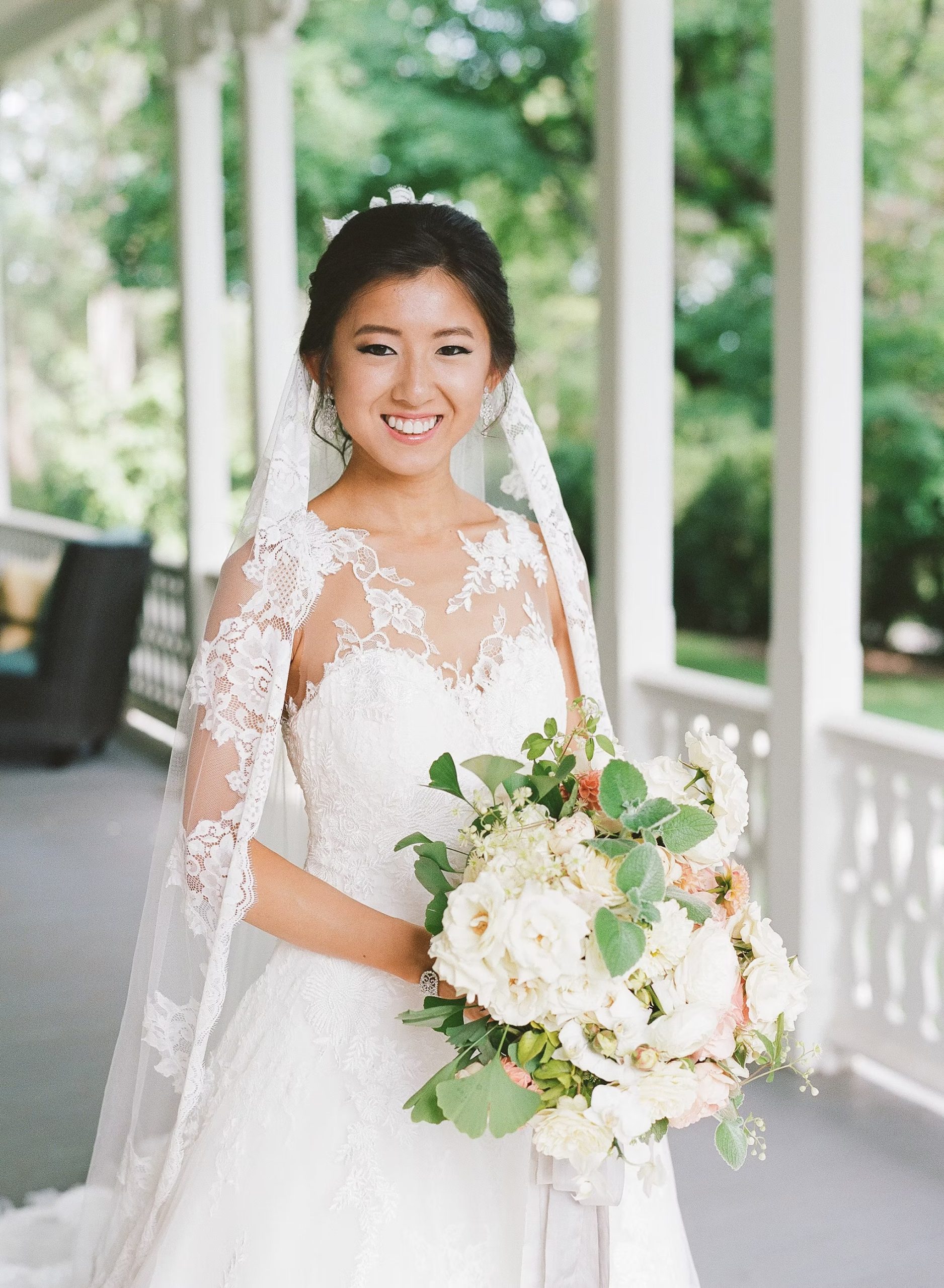Bride in lace wedding dress with illusion neckline and veil, wearing updo hairstyle, holding white and greenery bouquet