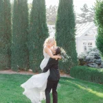 Groom lifts bride in elegant garden setting with tall cypress trees and white gazebo in background