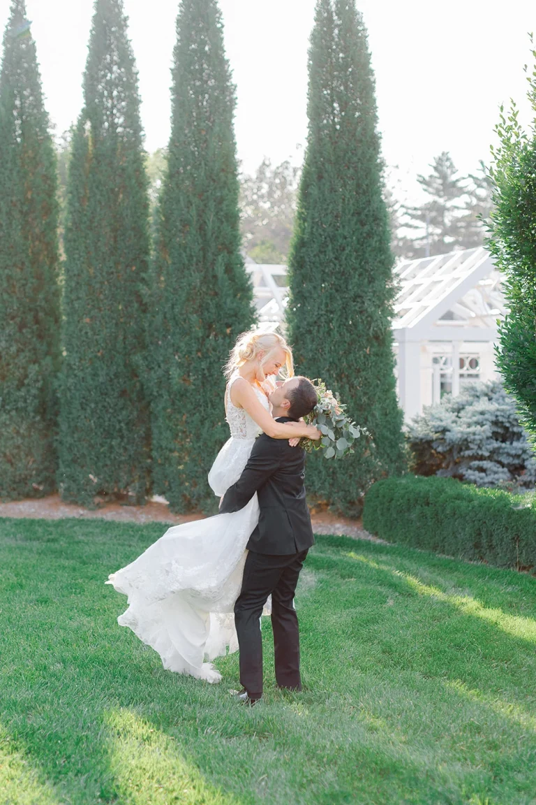 Groom lifts bride in elegant garden setting with tall cypress trees and white gazebo in background