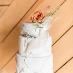 Three-tier white marble-textured wedding cake with clear acrylic sail detail, topped with a terracotta rose and greenery