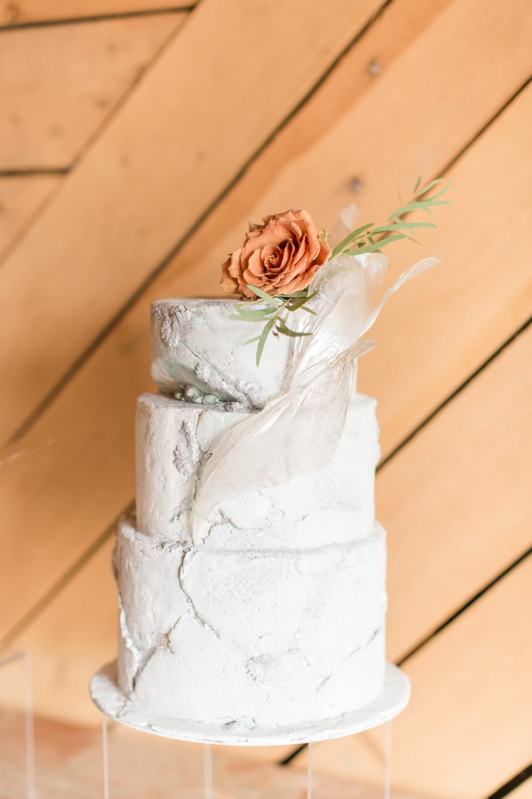 Three-tier white marble-textured wedding cake with clear acrylic sail detail, topped with a terracotta rose and greenery