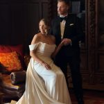 Bride in off-shoulder white gown seated beside groom in black tuxedo in elegant dark interior setting