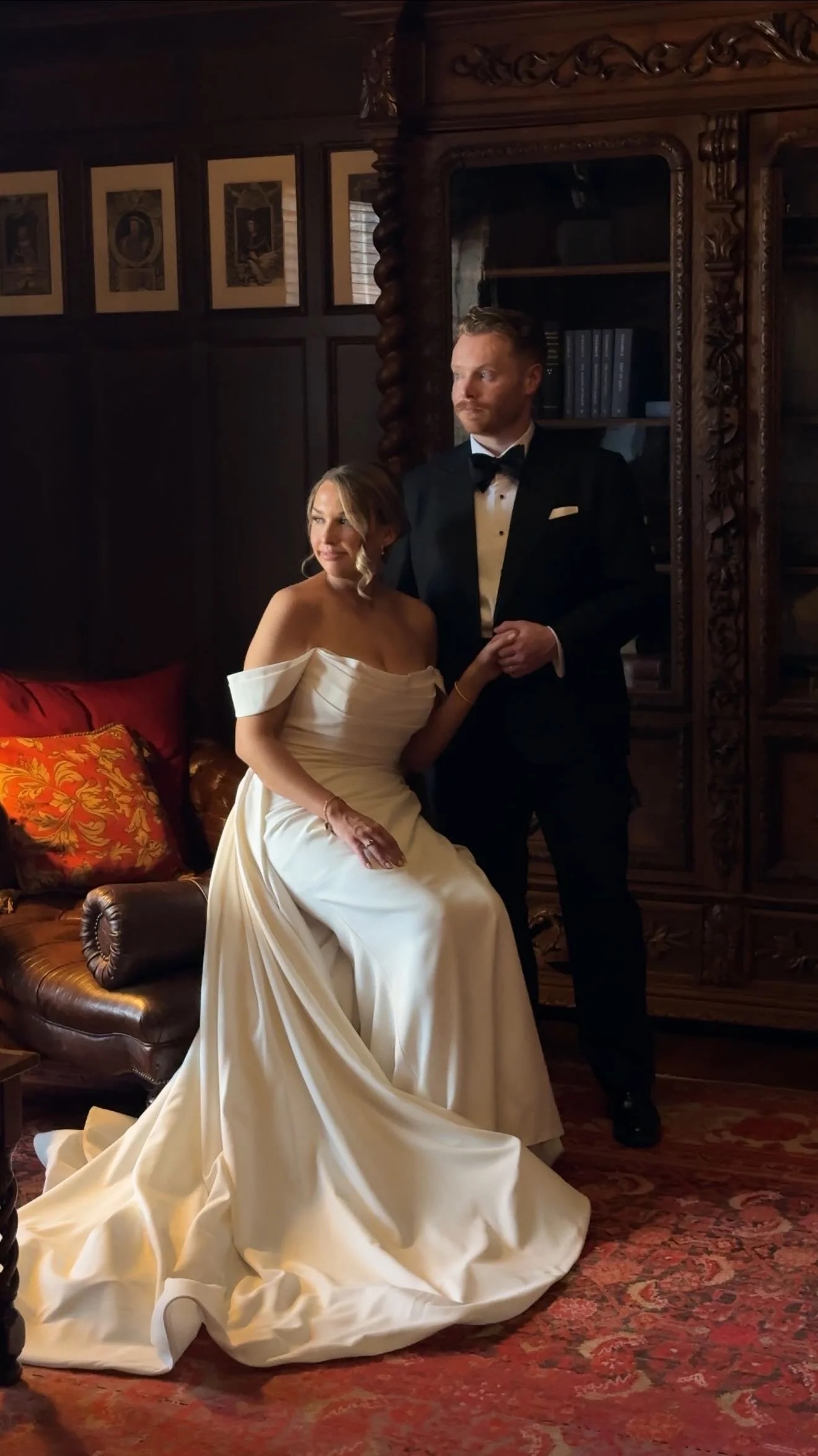 Bride in off-shoulder white gown seated beside groom in black tuxedo in elegant dark interior setting