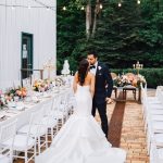 Bride in mermaid gown and groom in navy suit kiss between reception tables under string lights