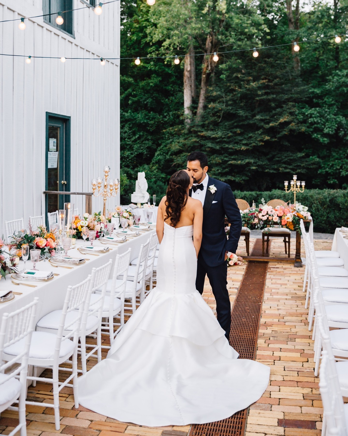 Bride in mermaid gown and groom in navy suit kiss between reception tables under string lights