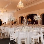 Wide view of elegant ballroom reception with white tables, chandeliers, and tall floral arrangements