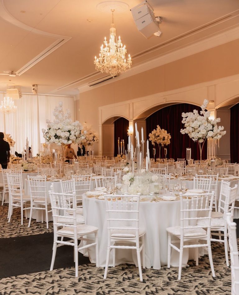 Wide view of elegant ballroom reception with white tables, chandeliers, and tall floral arrangements
