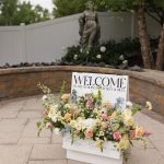 Welcome sign with floral arrangement in pink and peach tones displayed on patio near fountain