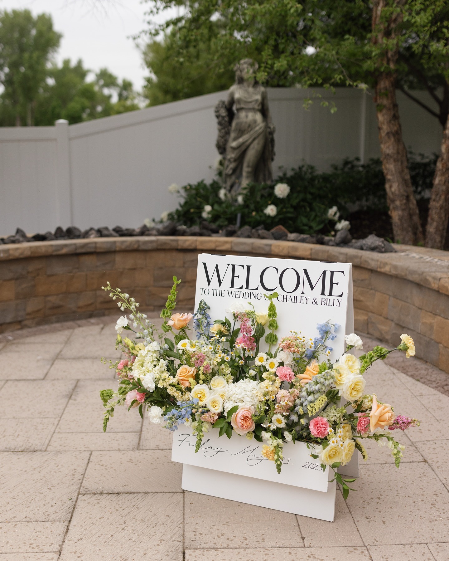 Welcome sign with floral arrangement in pink and peach tones displayed on patio near fountain