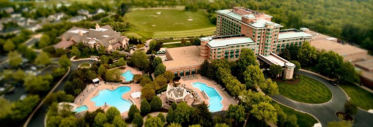 Aerial view of resort property with swimming pools, golf course, and multi-story hotel buildings surrounded by trees