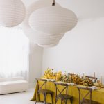 Airy white reception space showcasing mustard table draping, yellow and amber floral centerpiece, and sculptural paper lanterns
