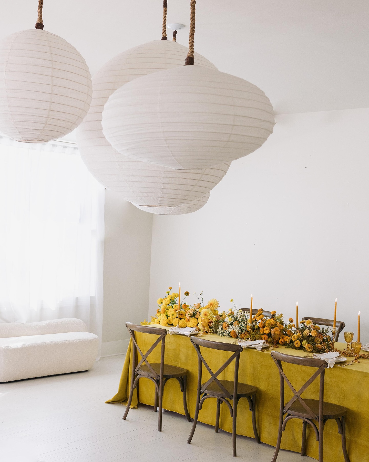 Airy white reception space showcasing mustard table draping, yellow and amber floral centerpiece, and sculptural paper lanterns