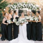 Bride and bridesmaids in black gowns holding white bouquets in front of floral mantle arch