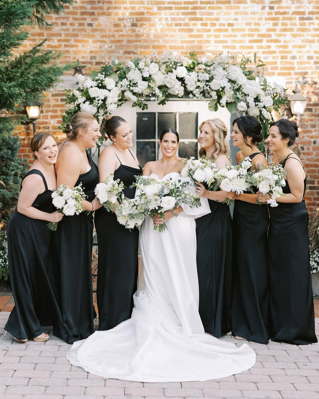 Bride and bridesmaids in black gowns holding white bouquets in front of floral mantle arch