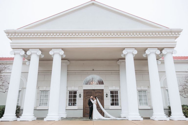 Bride and groom embracing under grand white colonial portico with Ionic columns and triangular pediment