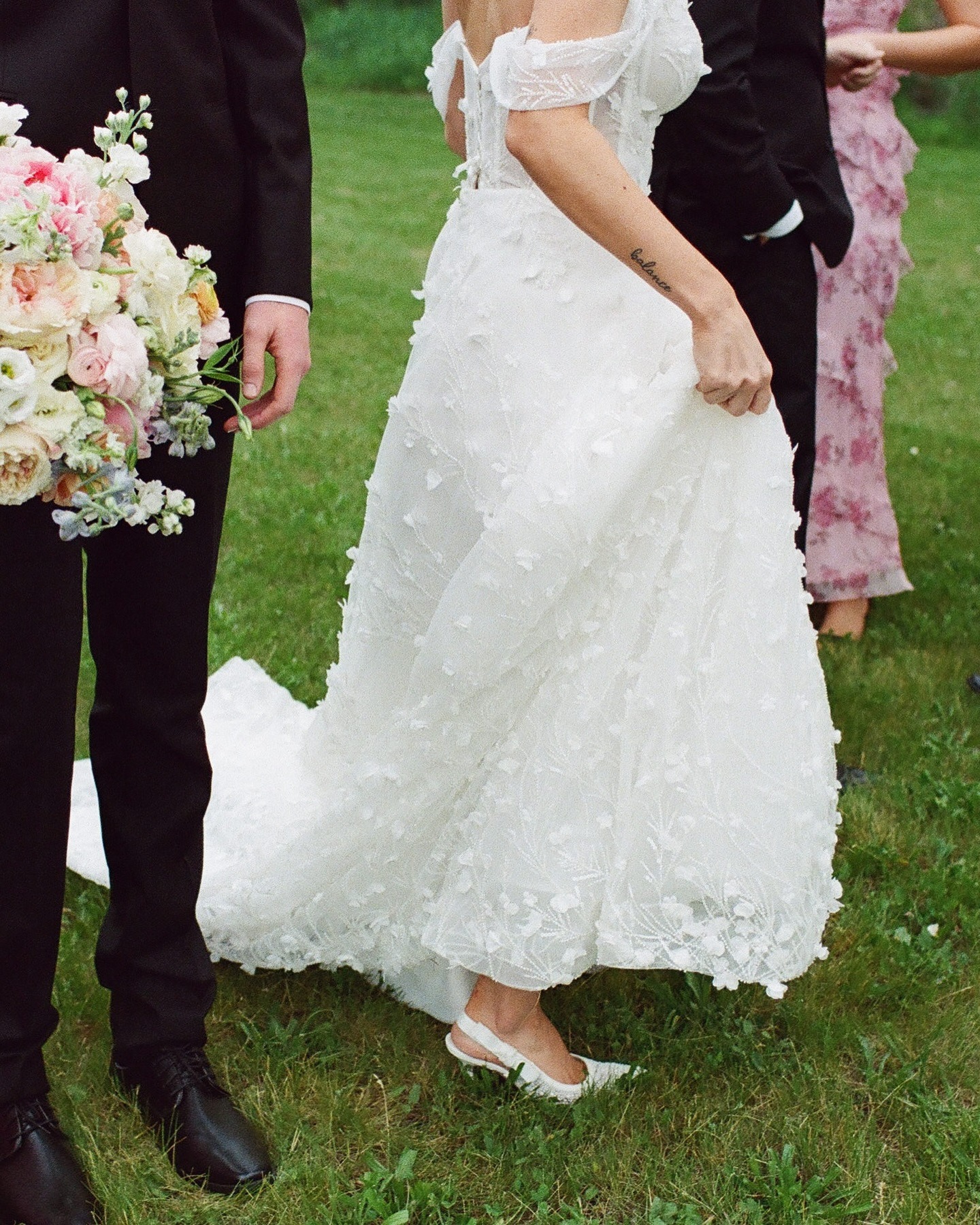Close-up of bride's textured white gown and groom's black suit as they hold hands on grass