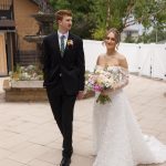 Newlyweds walk hand-in-hand on courtyard patio, bride carrying pastel bouquet
