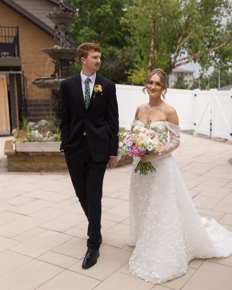 Newlyweds walk hand-in-hand on courtyard patio, bride carrying pastel bouquet