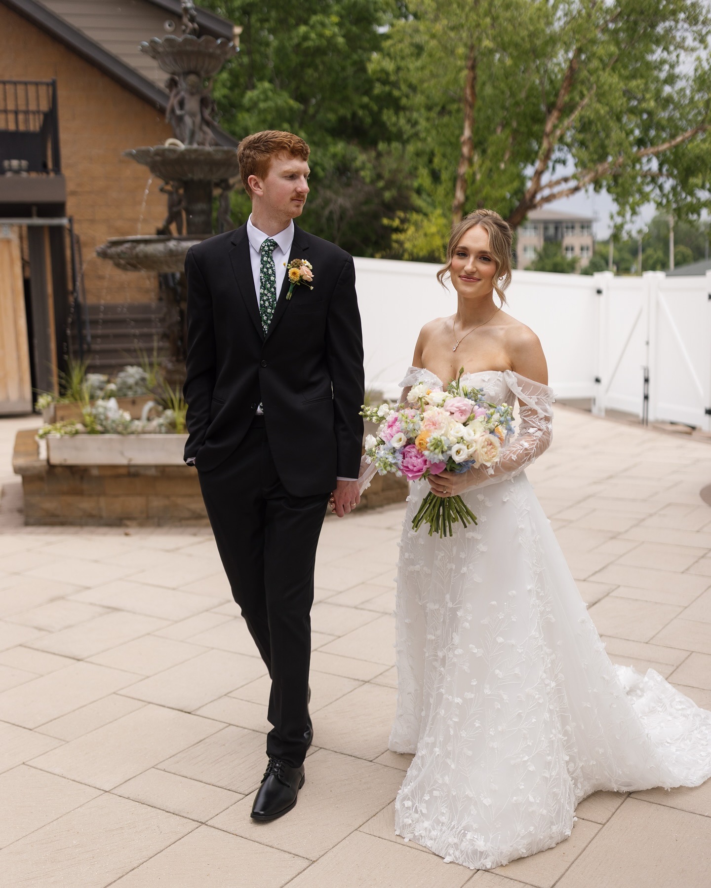 Newlyweds walk hand-in-hand on courtyard patio, bride carrying pastel bouquet
