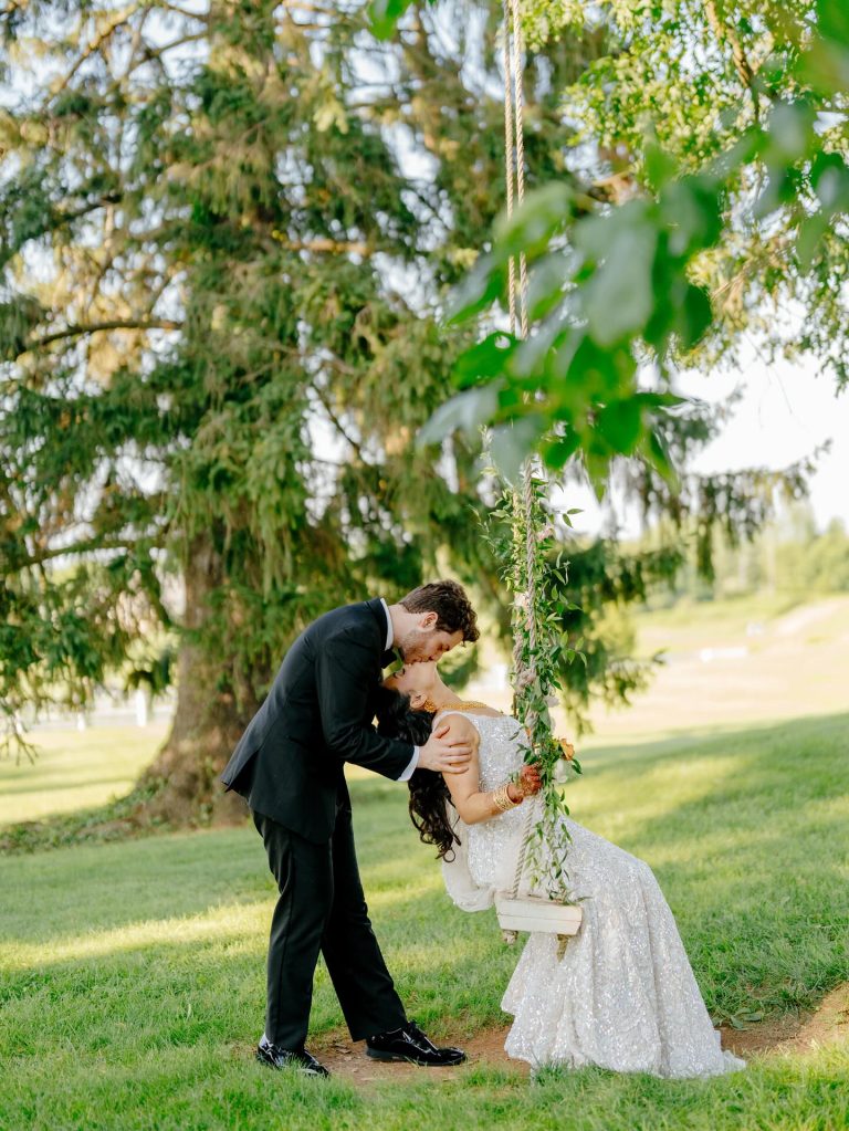 Groom leaning in to kiss bride seated on swing decorated with greenery beneath weeping willow tree