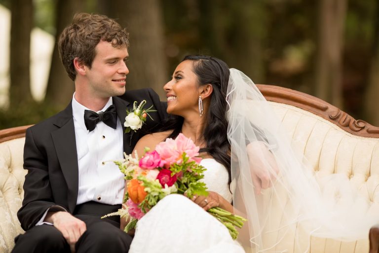 Bride with cascading waves and chandelier earrings smiling at groom on elegant outdoor settee
