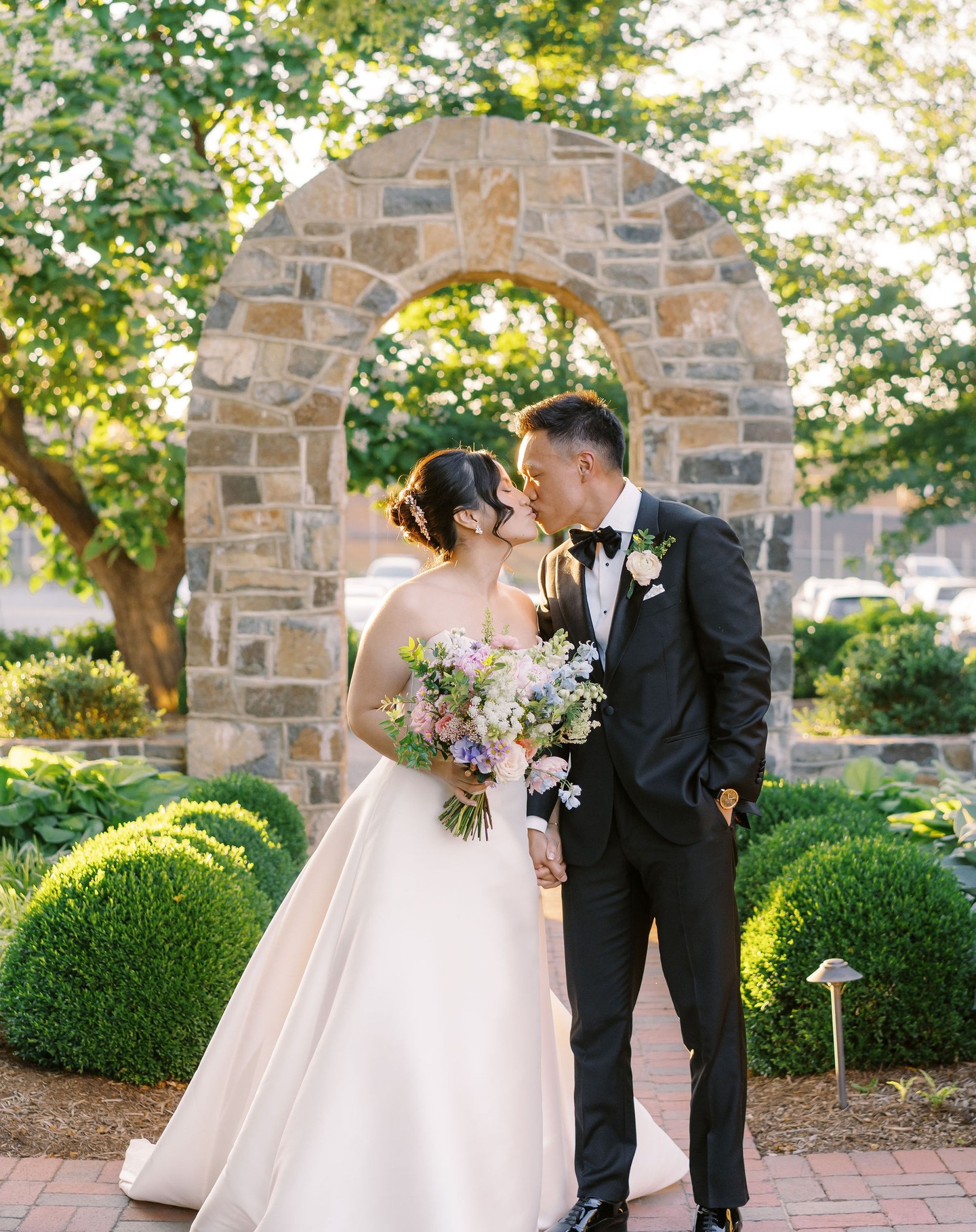 Newlyweds kissing in garden with stone archway and boxwood hedges