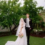 Newlyweds posing in garden setting with bride holding pastel bouquet beneath veil