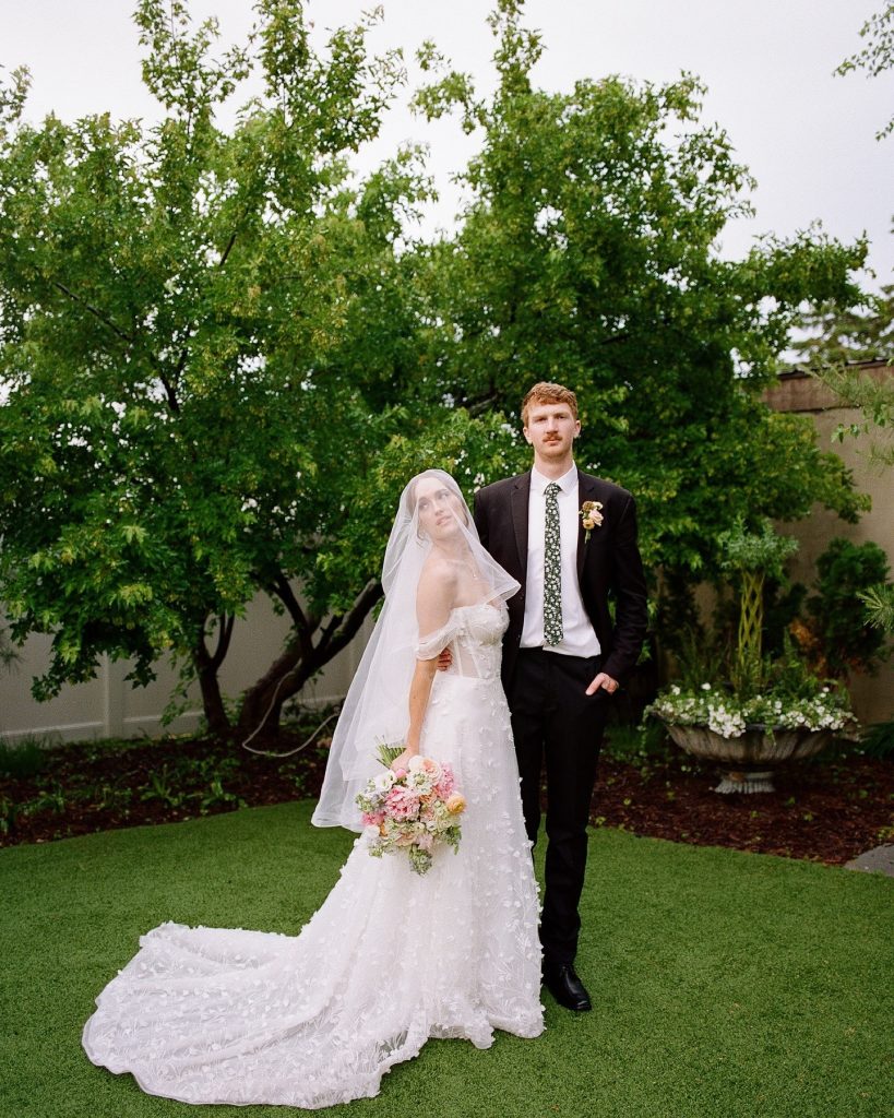 Newlyweds posing in garden setting with bride holding pastel bouquet beneath veil