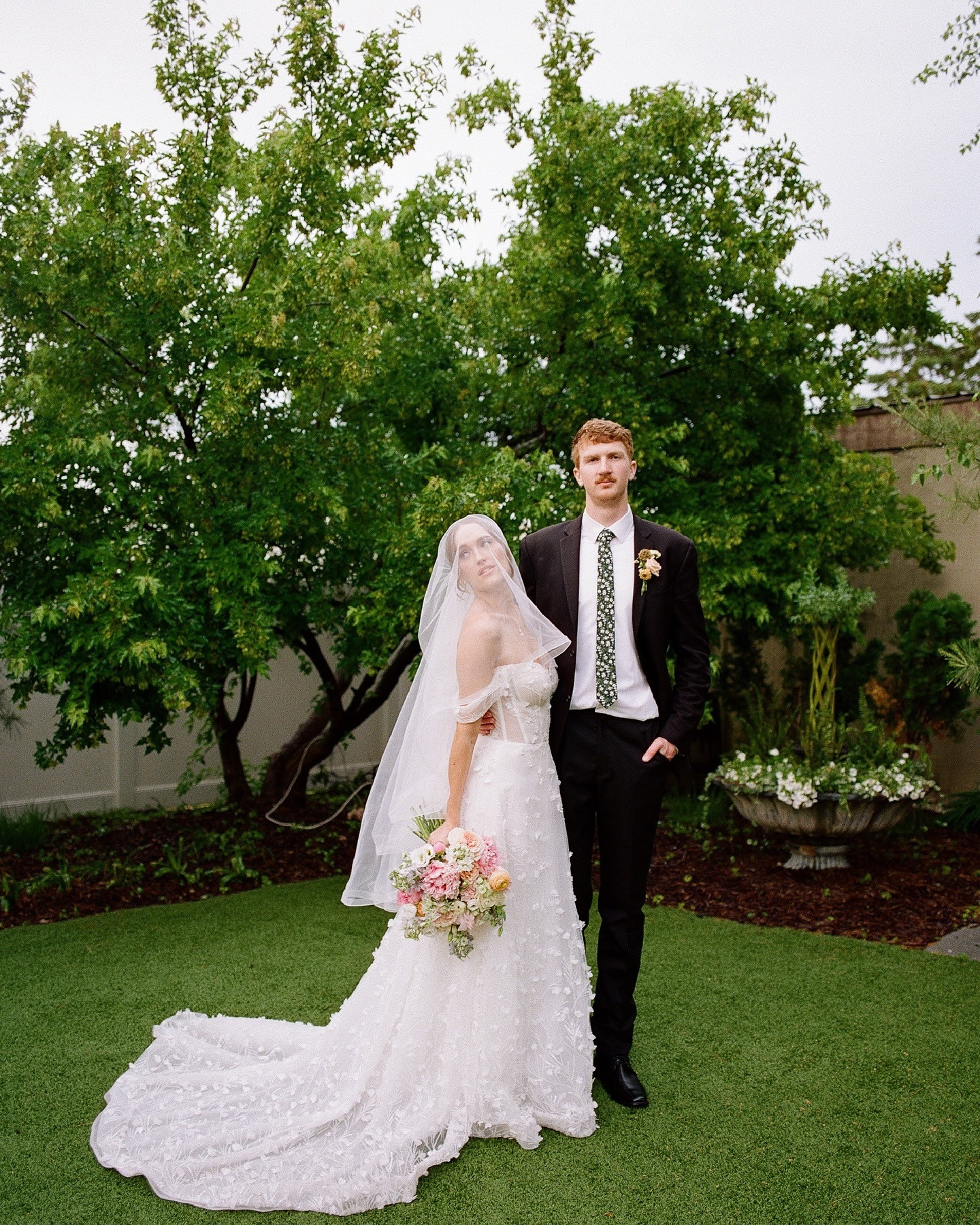Newlyweds posing in garden setting with bride holding pastel bouquet beneath veil