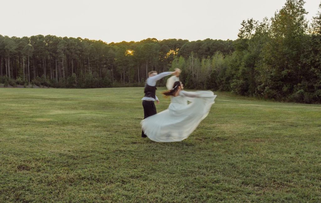 Newlyweds spinning together at sunset in open field with bride's flowing dress and forest backdrop