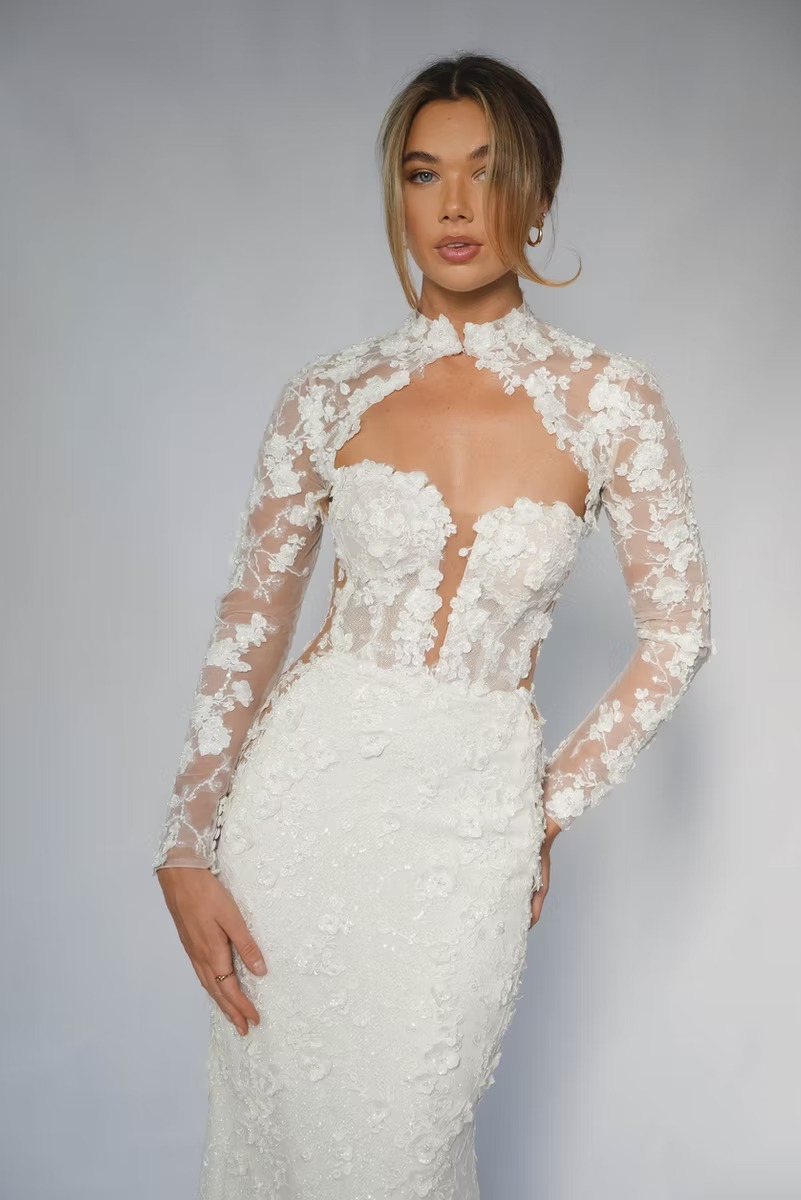 Fitted wedding dress with sheer long sleeves and high neckline featuring white floral lace appliqués