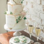 Three-tier white wedding cake with gold drip detail and ivy leaves, displayed with champagne flutes and green swirl meringues