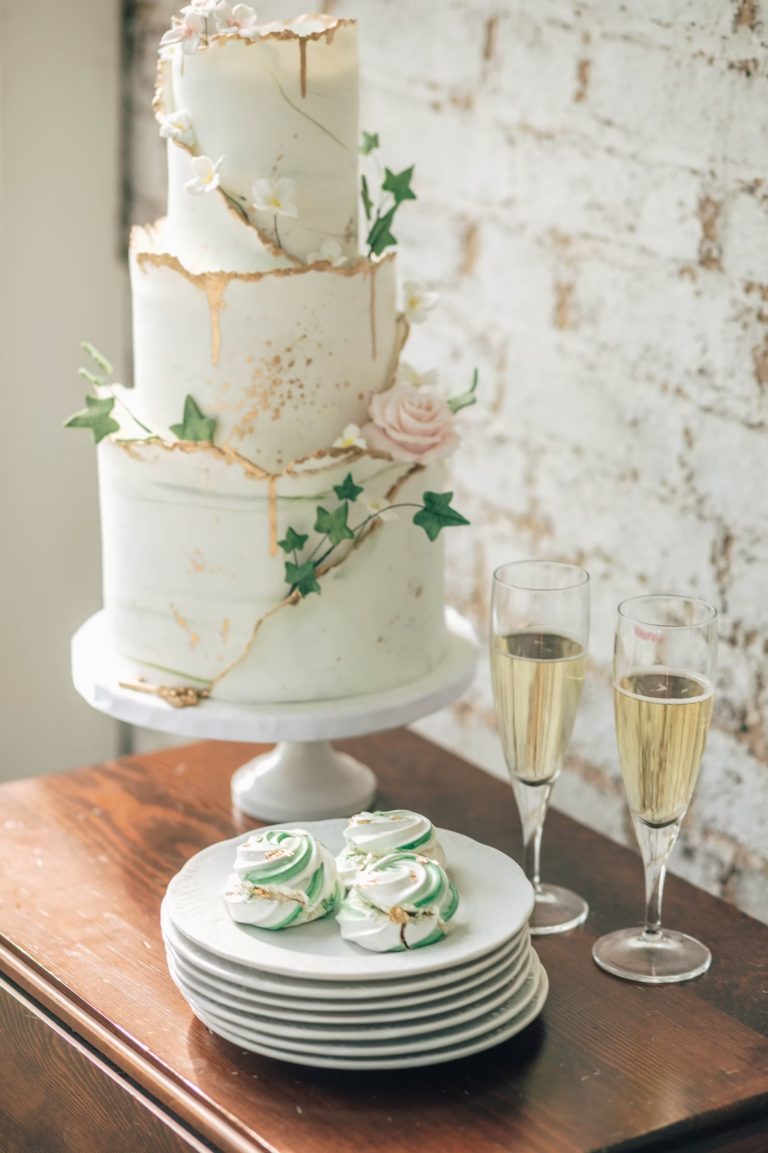 Three-tier white wedding cake with gold drip detail and ivy leaves, displayed with champagne flutes and green swirl meringues