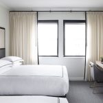 Modern hotel guest room with black-framed bed, neutral tones, large windows, and minimalist desk area