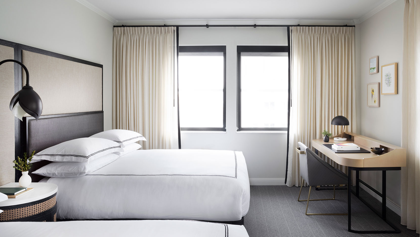 Modern hotel guest room with black-framed bed, neutral tones, large windows, and minimalist desk area