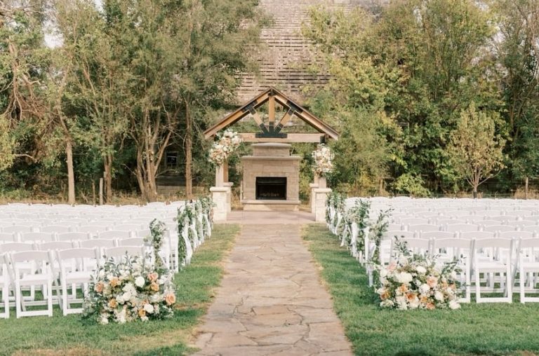 Outdoor ceremony space at The Barn at Ackerhurst Dairy Farm featuring white chairs, floral aisle decor, and stone fireplace