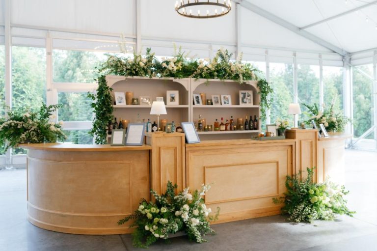 Wooden bar station decorated with lush greenery and white florals in a bright tent venue with glass walls
