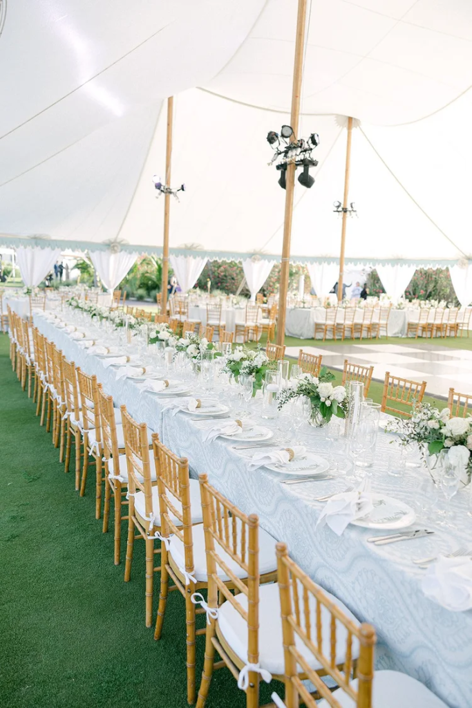 Long reception table with gold chiavari chairs, white linens, and greenery centerpieces under sailcloth tent