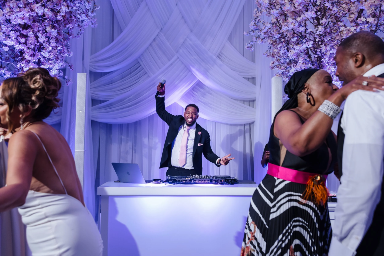 Wedding DJ in tuxedo energizing guests at elegant reception with purple uplighting and floral decor