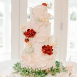 Four-tier wedding cake with blush pink frosting, gold accents, decorated with red peonies and cream roses, surrounded by greenery and rose gold votives