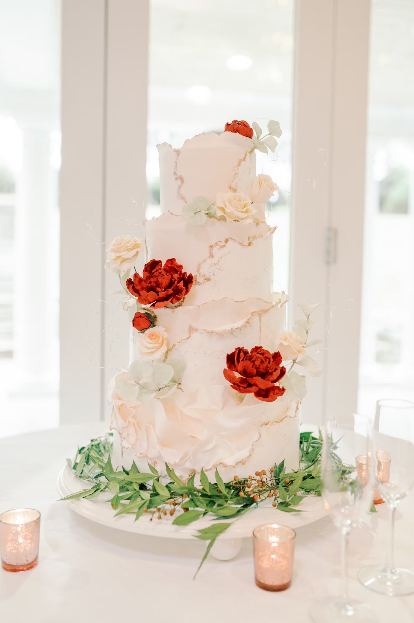 Four-tier wedding cake with blush pink frosting, gold accents, decorated with red peonies and cream roses, surrounded by greenery and rose gold votives